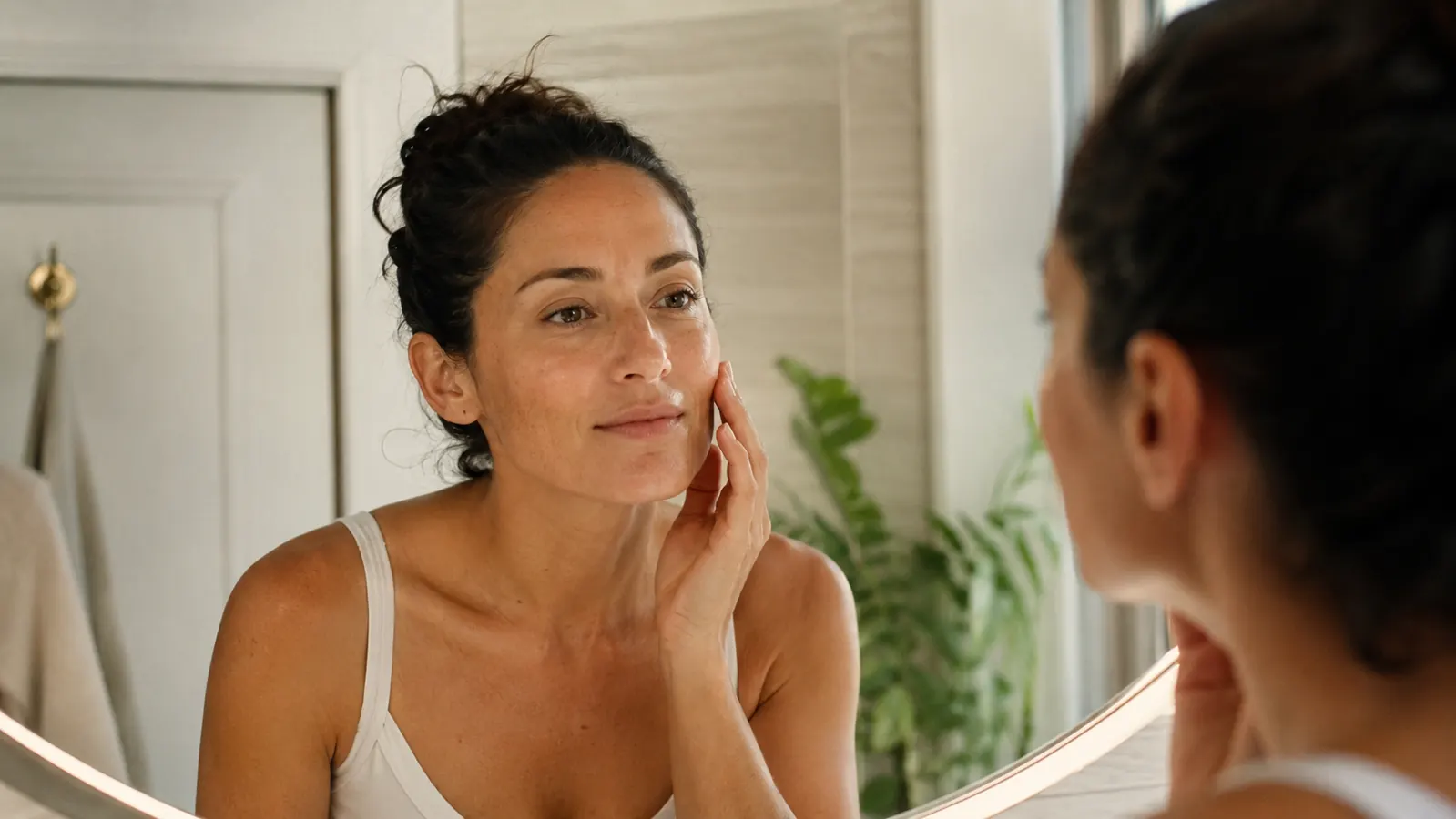 Woman touching her cheek in a bright bathroom mirror, skin looking calm and even without visible sunscreen cast