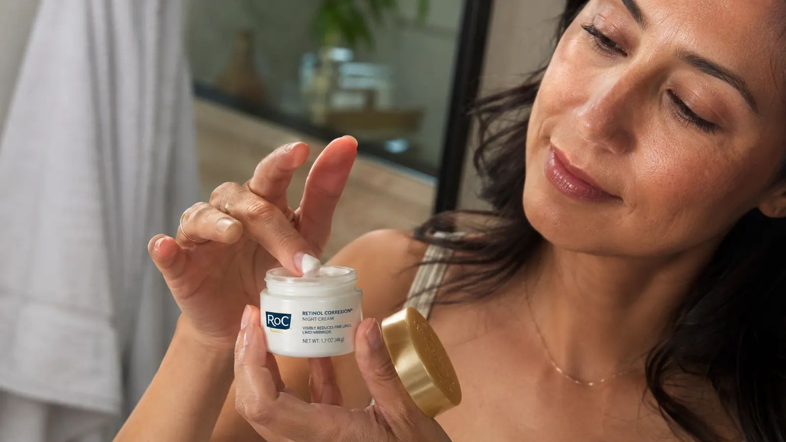 Hand holding a small jar of RoC Retinol Correxion open, finger mid-dip into the cream, bathroom mirror in background