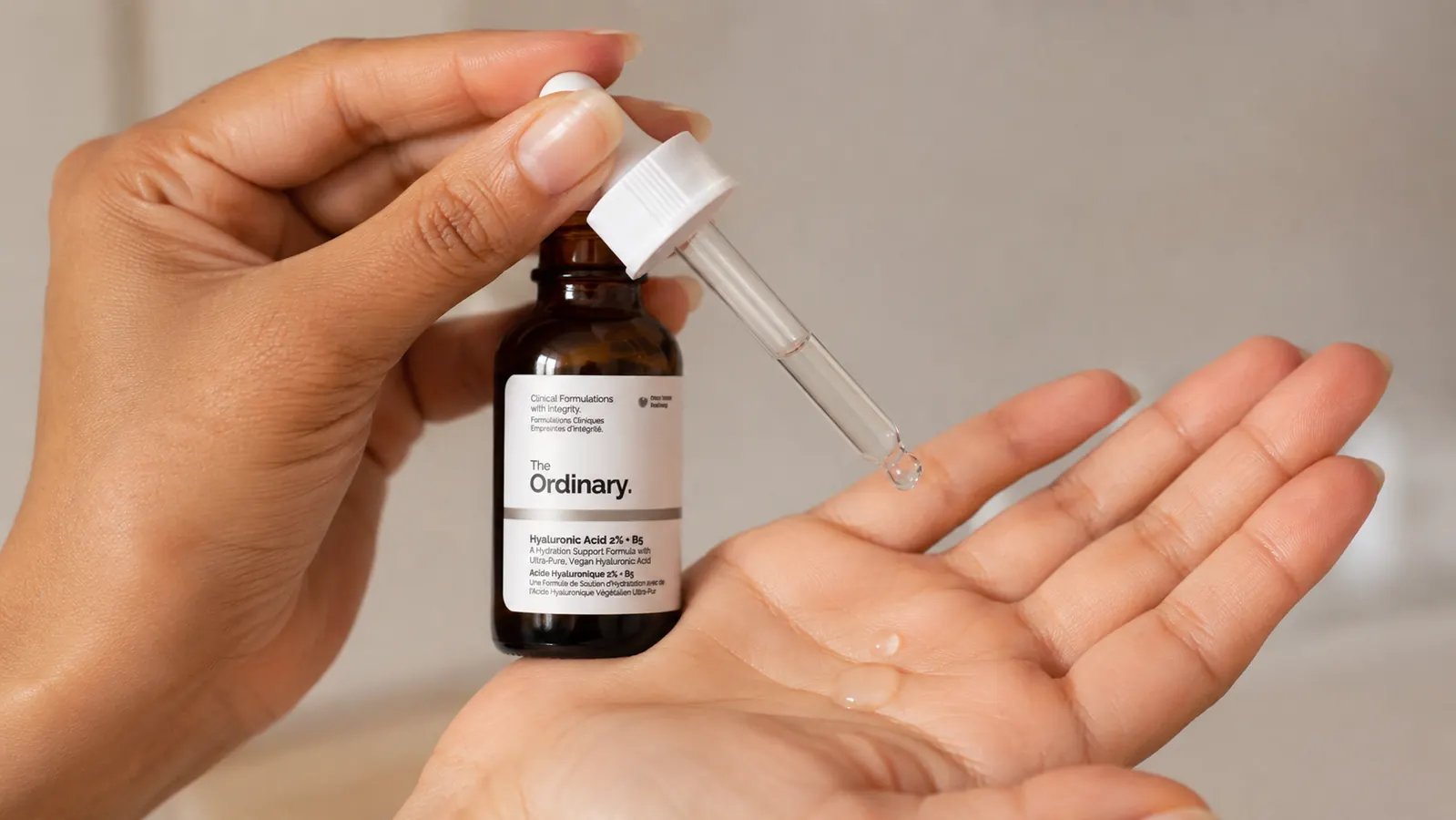 Hand holding The Ordinary Hyaluronic Acid dropper bottle over a palm with a few drops of clear serum